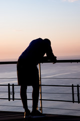 Man leaning on railing after a run while sun sets over the ocean