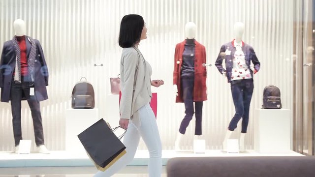 Young woman walks along a show window with bags and phone in her hand in the shopping mall. Side view dolly shot in slow motion.