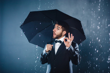 Cheerful handsome bearded young businessman posing with umbrella under the rain and showing OK...