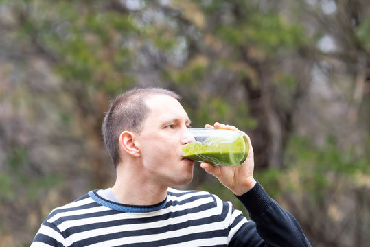 Young Man Standing Outside, Outdoors, Holding, Drinking From Glass Green Smoothie Made From Vegetables, Greens