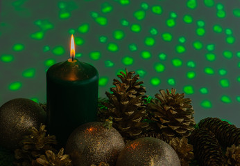 A single candle in the midst of gold pine cones on greenery with green lights in the background