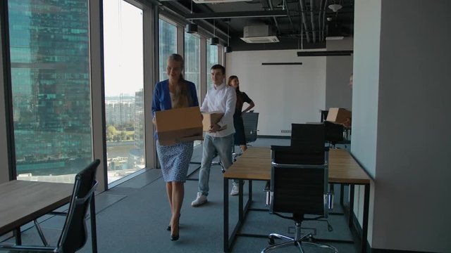 Female Boss and group of cheerful office workers walking through hallway towards the camera while moving into new office. Colleagues carrying cardboard boxes with belongings, computer. Wide shot.
