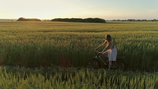 Cute young girl on bicycle in green field at sunset, drone shooting 4k