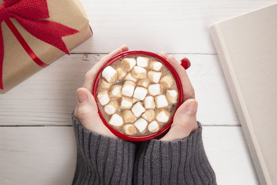 Person Holding a Hot Chocolate with Marshmallows on a Wooden Table with a Gift and a Novel to Read