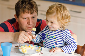 Adorable baby girl eating noodle, pasta macaroni. Father helping daughter learning to use spoon....