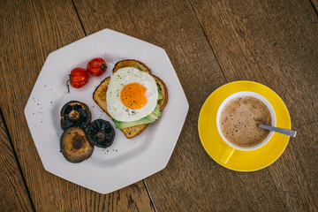 breakfast of fried egg on toast with mushrooms, avocado and tomato on wooden table.