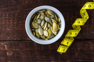 Peeled pumpkin seeds in a white saucer and measuring tape on a wooden background. Diet
