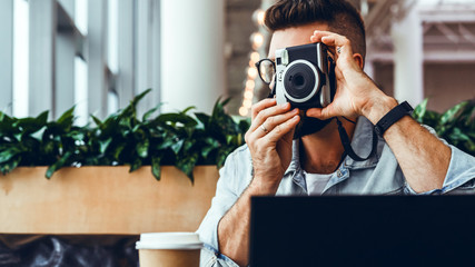 Young bearded man sits in coffee shop at table in front of laptop and takes instant photo on camera.