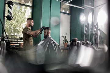 Young man with a beard sits in the chair at a barber shop. Barber dries mens hairs.