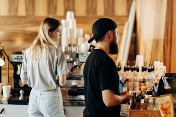 Two young stylish people,a thin blonde girl and a man with beard,wearing casual clotes,cook coffee in a modern coffee shop.