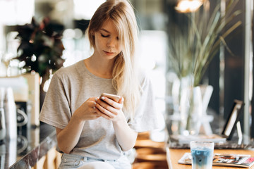 A cute thin blonde girl,dressed in casual style,drinks coffee and looks at her phone in a coffee shop.