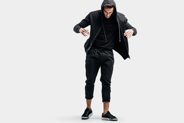 Dark-haired stylish dancer wearing black clothes and a hood makes stylized movements of street dance.