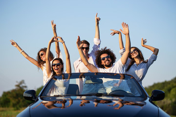 Happy young girls and guys in sunglasses are sitting in a black cabriolet on the road holding their...