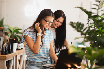 Two cute slim girls with long dark hair,wearing casual style,sit at the table and look attentively at the laptop screen in a cozy coffee shop.