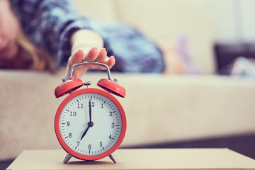 Young girl lies on the couch and stretches her hand to the red alarm clock to turn it off. Late...