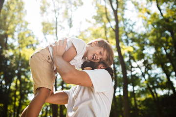 Dark-haired father with beard dressed in the white t-shirt is holding in the arms his blond son and smiling on a summer day in the park.