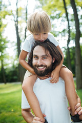 Father and his son walking in the forest. Boy dressed in a white t-shirt sitting on the shoulders of his father.