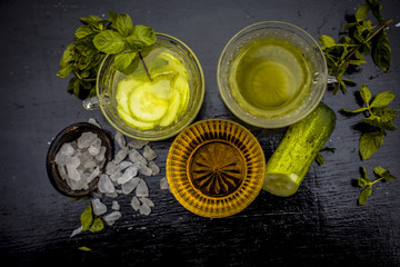 Fototapeta na wymiar Iced cucumber and mint tea on wooden surface in a transparent cup with slices of cucumber and raw cucumber,mint leaves,sugar and honey with green tea in a separate glass cup.