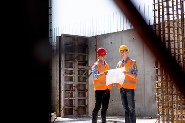 Structural engineer and construction manager in orange work vests and hard helmets explore construction documentation on the building site near the steel frames