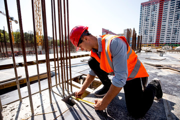 Civil engineer dressed in shirt, orange work vest and helmet measures the hole with a tape measure on the building site