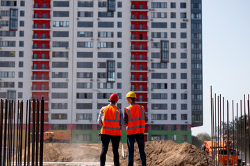 Two men dressed in orange work vests and helmets talk about  the construction process on the building site against the background of a multi-storey building