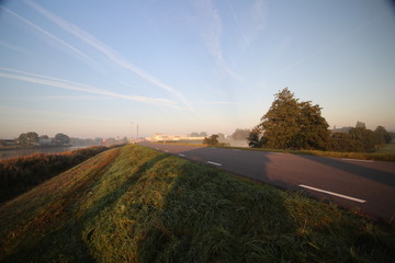 Sunrise with colored aircraft trails, fog on the meadows and dyke at River Hollandsche IJssel in the Netherlands at nieuwerkerk.