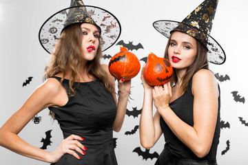 Two brunette girls in black dresses and witch`s hats  grimace and hold halloween pumpkins in their hands