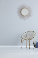 Gold mirror on grey empty wall above armchair in simple hall interior with copy space. Real photo