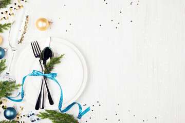 Christmas table setting with blue and golden balls, evergreens, blue ribbon, serpentine and sparkles on white wooden table. Overhead.