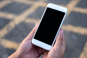 Mockup image of hand holding white mobile phone with blank black screen with street background