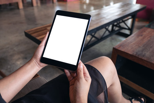 Mockup image of a woman sitting and holding black tablet pc with blank white desktop screen in wooden cafe