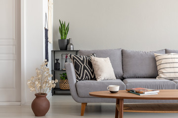 Flowers next to grey couch with pillows in minimal living room interior with wooden table. Real photo