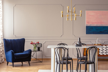 Dark blue armchair in a dining room interior with a table, chairs and golden lamp. Real photo. Empty wall, place your graphic