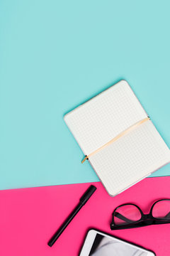Open white Notepad, glasses, smartphone, pens on colored blue, pink background
