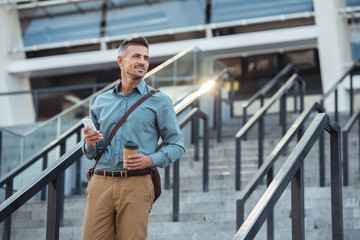 smiling middle aged businessman with coffee to go and smartphone looking away on stairs