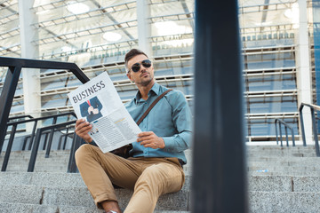handsome man in sunglasses holding business newspaper and looking away while sitting on stairs