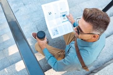 high angle view of man in sunglasses holding paper cup while sitting on stairs and reading newspaper