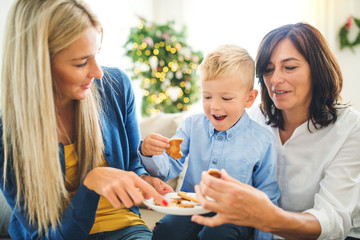 A small boy with mother and grandmother eating biscuits at home at Christmas time. - obrazy, fototapety, plakaty