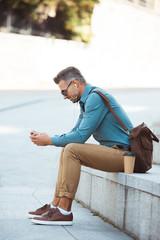 side view of man in earphones and sunglasses sitting on stairs and using smartphone