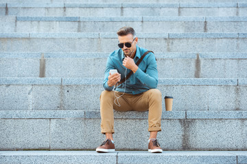 Fototapeta premium stylish middle aged man in earphones sitting on stairs and using smartphone