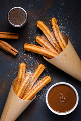 Churros with sugar powder with chocolate sauce dip and cinnamon sticks on dark background top view
