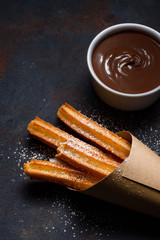 Churros in paper bag with sugar and chocolate sauce on dark black background