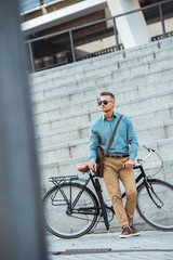 Fototapeta premium handsome middle aged businessman in sunglasses holding umbrella while standing near bicycle on street