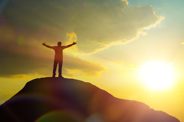 A man at the top of the mountain, raising his hands up against the backdrop of the glare of the sun. Concept of success and achievements, business career ladder, victory over competitors