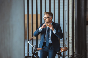 smiling middle aged businessman in eyeglasses sitting on bike and holding hamburger