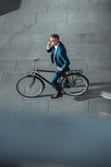 Fototapeta na wymiar high angle view of businessman in formal wear talking by smartphone while standing with bicycle on street