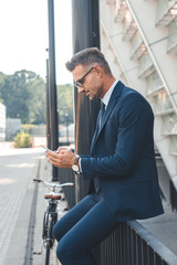 side view of businessman in suit and eyeglasses using smartphone on street