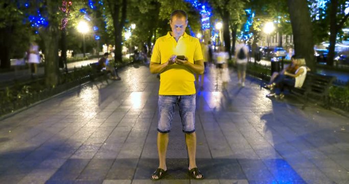 Timelapse Of Man Standing Still With Smartphone On Crowded Walkway Park Boulevard At Summer Night. People Moving Fast. Concept Of Flowing Time And Technologies