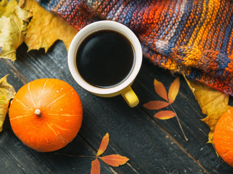 A mug of hot coffee, a warm scarf and a book on a dark wooden table strewn with autumn leaves