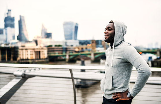 Black Man Runner With Smart Watch Standing On The Bridge In A City, Resting.